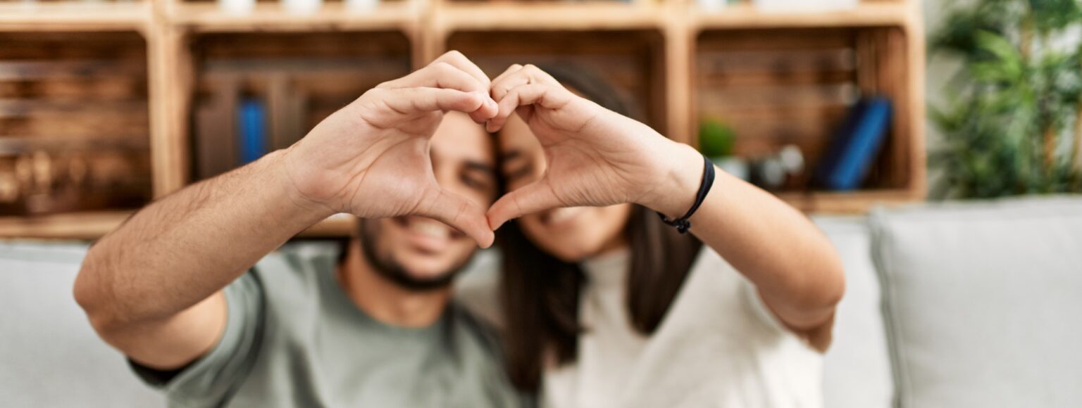 Young latin couple smiling happy doing heart symbol with hands at home.