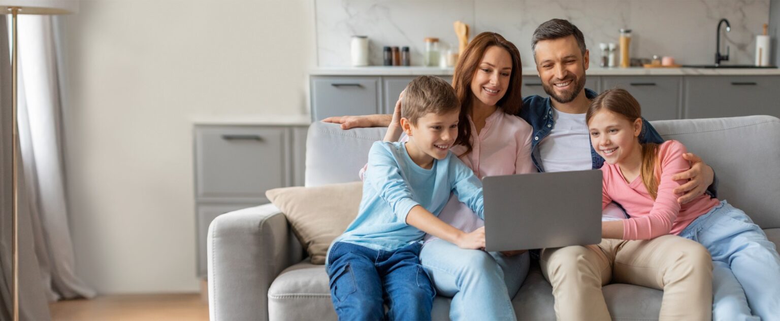 A family of four sits comfortably on a couch in their living room, watching a movie on a laptop. They share smiles and laughter, creating a cozy atmosphere in their home.