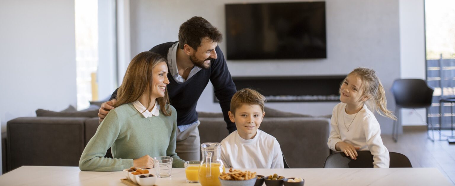 Young happy family talking while having breakfast at dining table at apartment